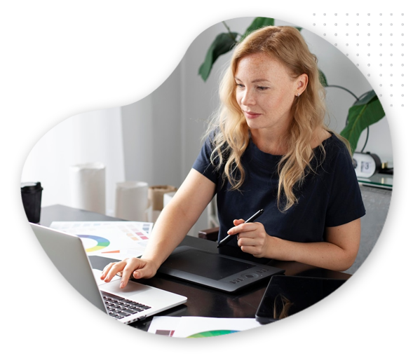 Blog-branding-img A woman with blonde hair sits at a desk using a laptop and a digital drawing tablet, working on blog branding, surrounded by colorful charts and papers, with plants in the background.