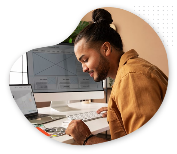 A person with long hair in a bun, wearing a brown shirt, sits at a desk working on web design wireframes on the computer, with a laptop nearby, holding a pen and color swatches—perfect inspiration for your next blog post.