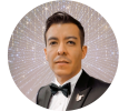 A man in a tuxedo and bow tie is smiling slightly. He is in front of a sparkling, starburst-like background, giving the image a glamorous and radiant effect.