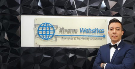 Robert Diaz - CEO of Xtreme Websites, stands with arms crossed before a black geometric wall and a sign that reads Xtreme Websites Branding & Marketing Solutions, a leader among top B2B companies, featuring a blue globe logo.