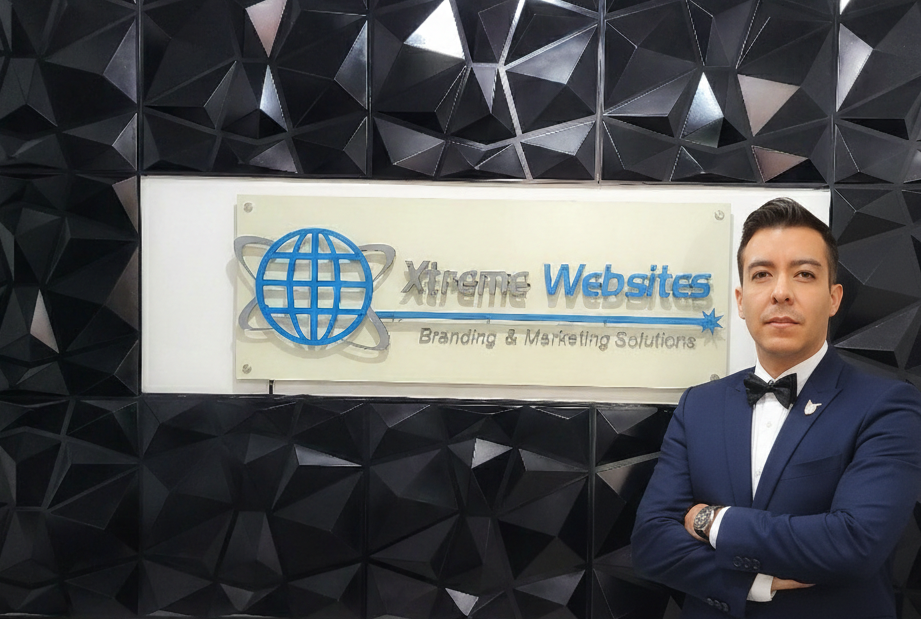 Robert Diaz - CEO of Xtreme Websites, stands with arms crossed before a black geometric wall and a sign that reads Xtreme Websites Branding & Marketing Solutions, a leader among top B2B companies, featuring a blue globe logo.