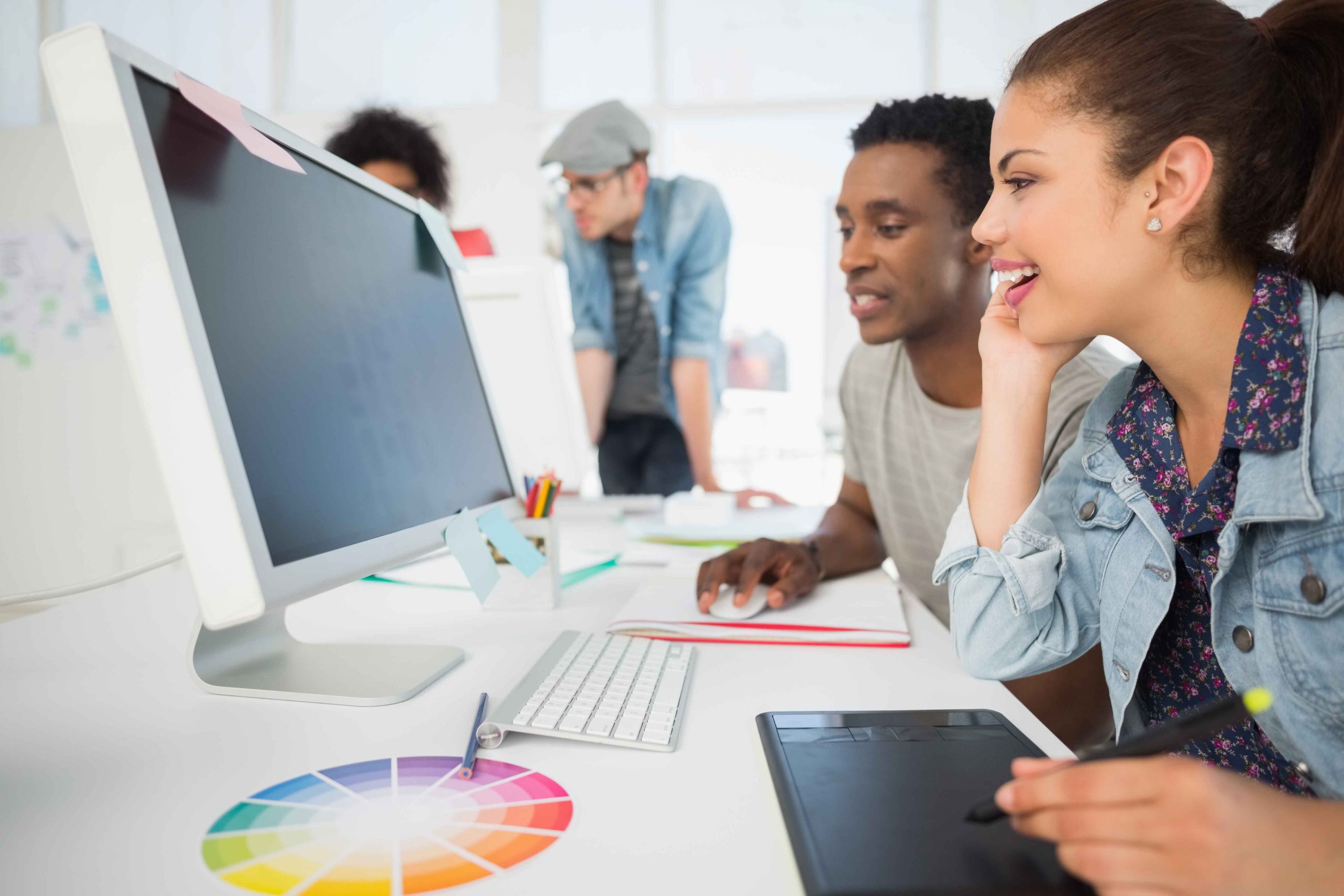 multimedia-design-3 Two people sit at a desk using a computer and digital drawing tablet, smiling and collaborating. In the background, two others stand and observe. A color wheel is visible on the desk, suggesting a creative project.